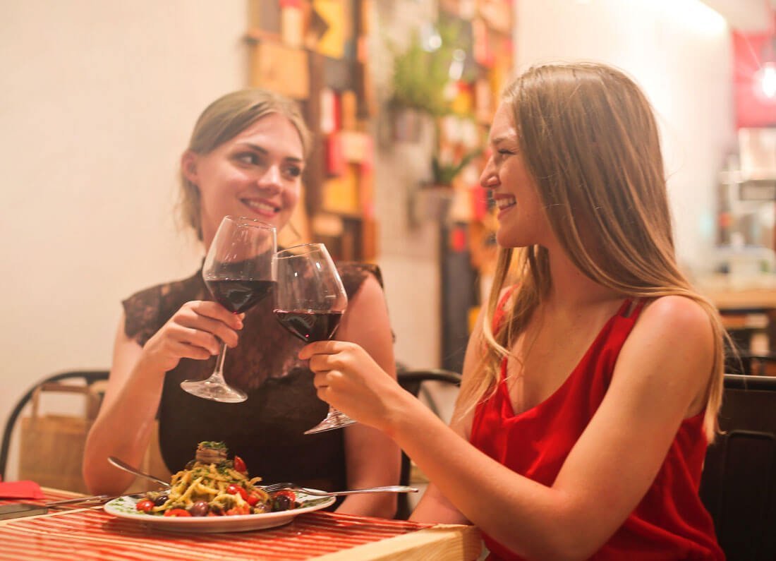 Two women clink wine glasses while enjoying a shared salad in a cozy restaurant bar/eatery scene