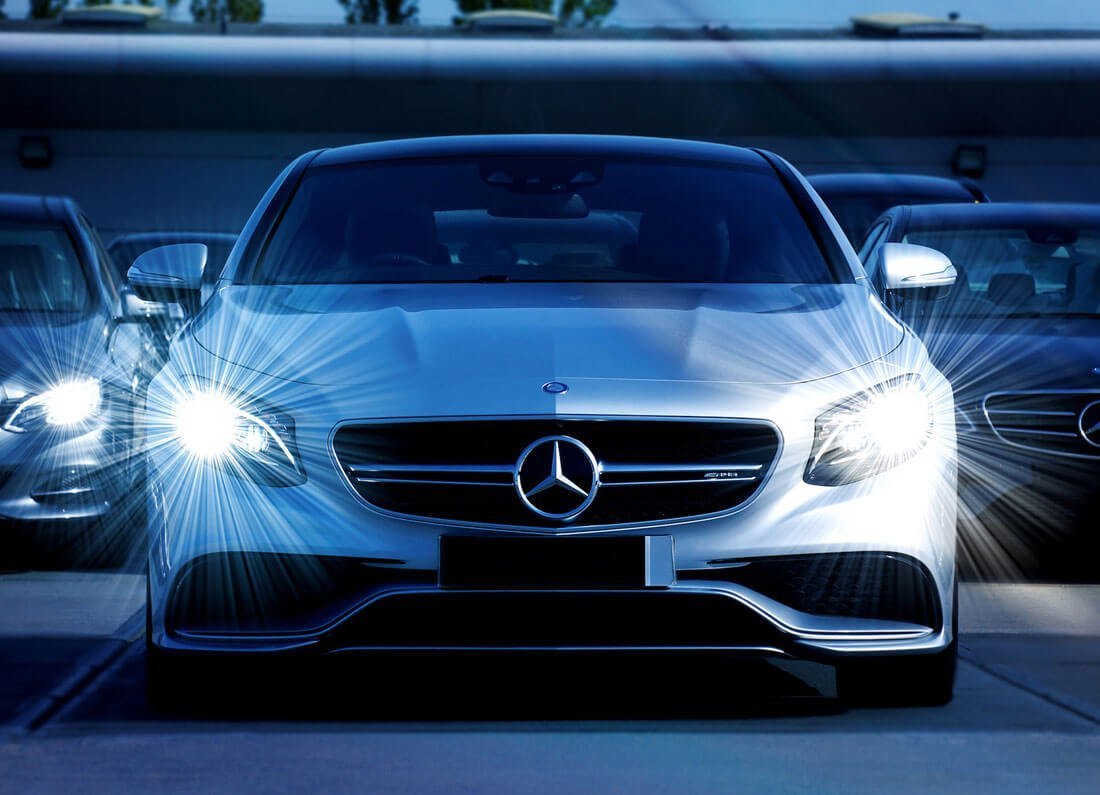marcedes-bens - Appalachian Highlands Farmers Magazine Front view of a silver Mercedes-Benz car with its headlights on, parked in a lot alongside other vehicles.
