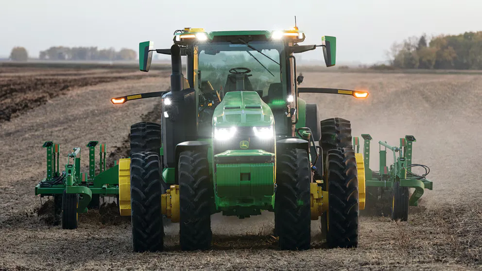 Front view of a green tractor pulling yellow tillage equipment across a dusty, bare field.