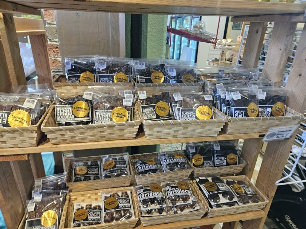 Assorted chocolate bars in clear bags on a wooden shelf display in a shop, labeled Cocoa Bean & Butter.