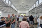 Busy indoor market or fair in a large white warehouse-like hall, people browsing tables of goods at vendor booths.