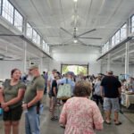 Busy indoor market or fair in a large white warehouse-like hall, people browsing tables of goods at vendor booths.