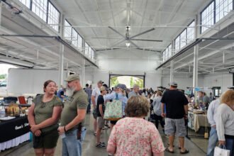 Busy indoor market or fair in a large white warehouse-like hall, people browsing tables of goods at vendor booths.