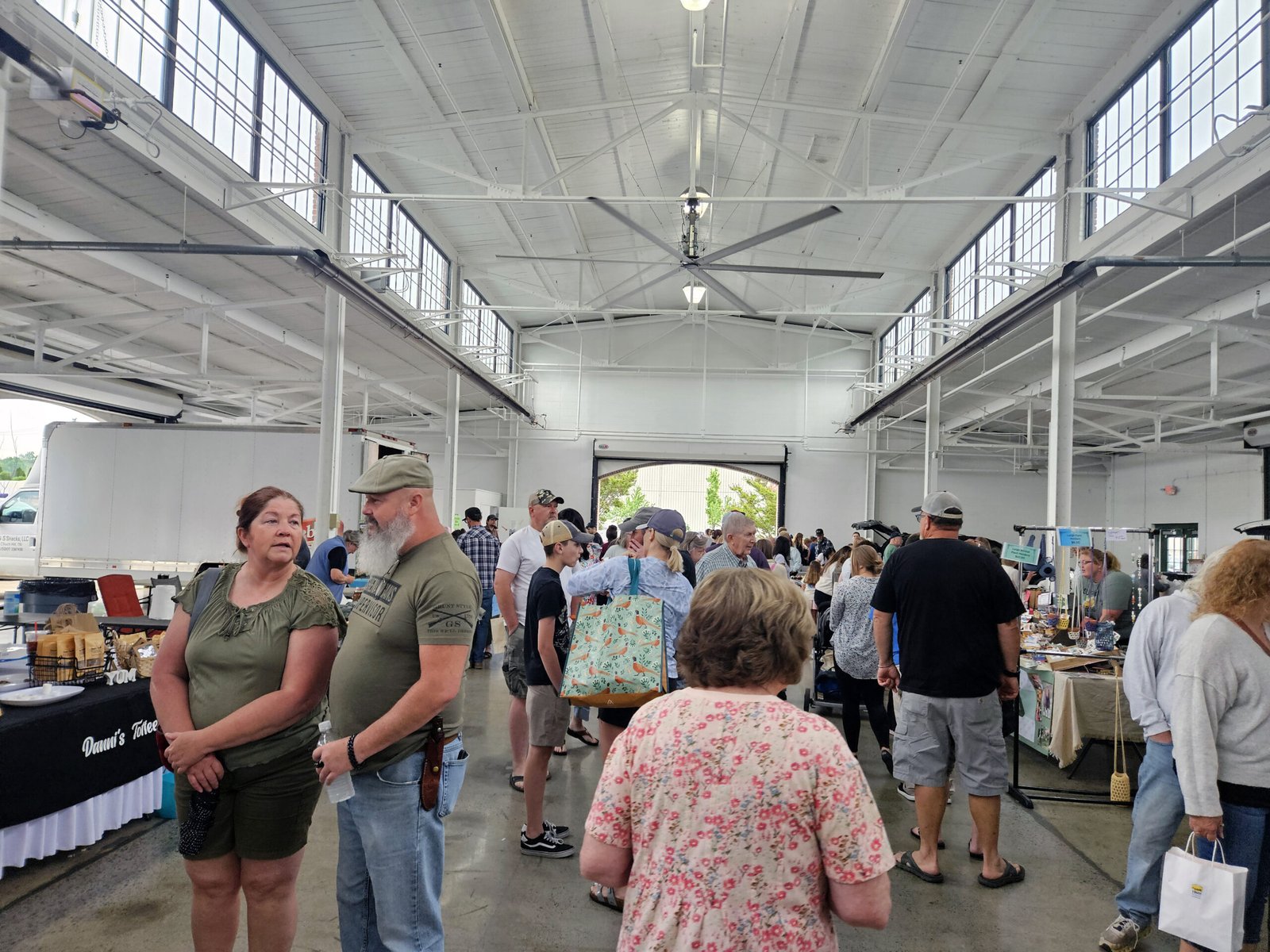 Busy indoor market or fair in a large white warehouse-like hall, people browsing tables of goods at vendor booths.
