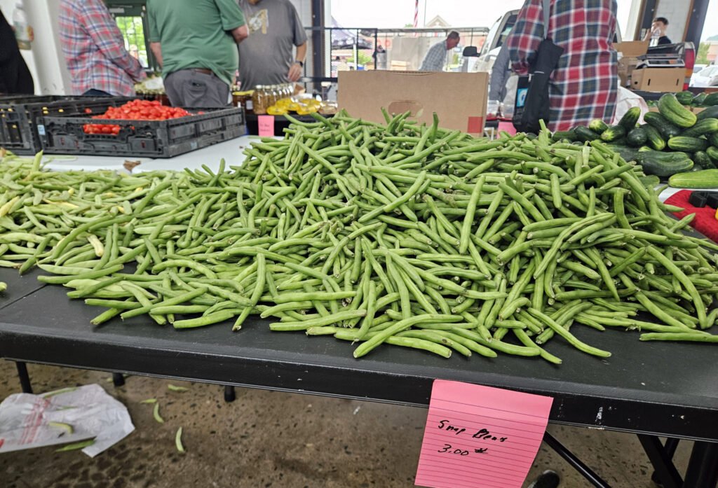 Pile of fresh green beans heaped on a market table, with vendors and produce in the background.