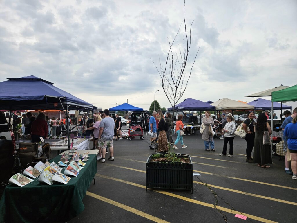 Outdoor street market with vendor stalls under blue, green, and beige canopies; shoppers browse tables of crafts beneath a cloudy sky.