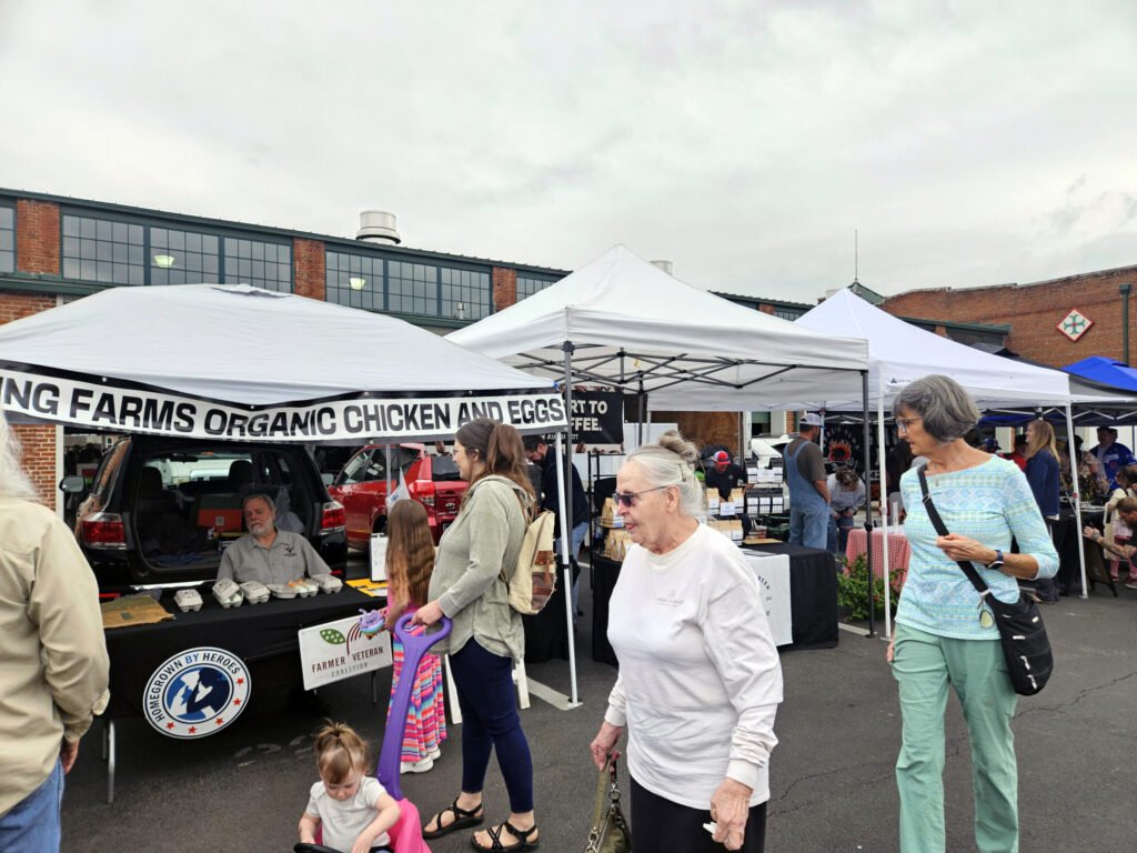 Outdoor market scene with white tents and a banner that reads 'FARMS ORGANIC CHICKEN AND EGGS', people browsing stalls and cars in the background.