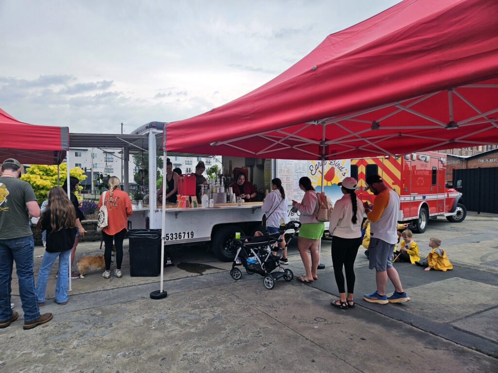 People gathered at an outdoor market under red tents, with a serving counter and a stroller nearby. A red emergency vehicle is visible in the background.