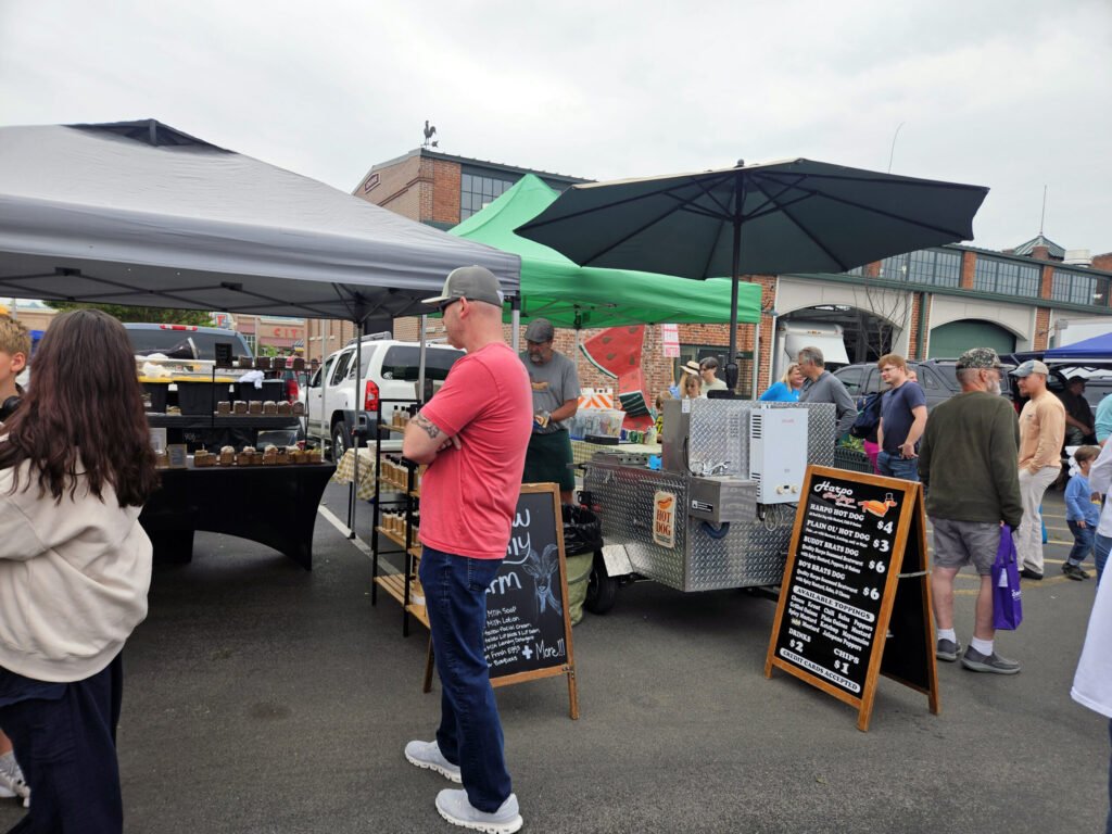 People browse an outdoor market with gray and green tents and vendor stalls in a parking lot, brick building in the background.
