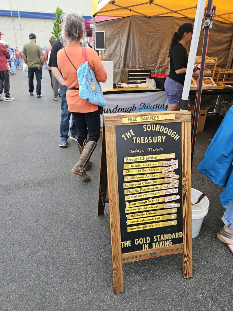 Wooden signboard for The Sourdough Treasury with 'Free Samples' and a flavor list at a market stall, shoppers in the background.