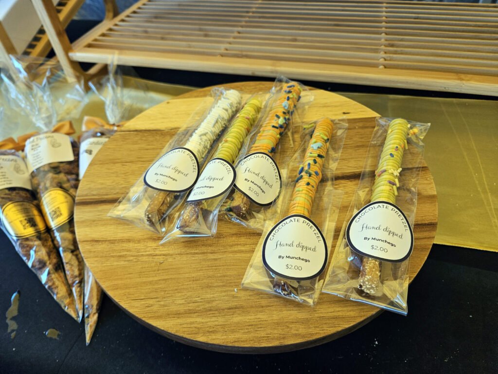Assorted hand-dipped chocolate pretzels in clear bags with circular labels on a wooden display table (