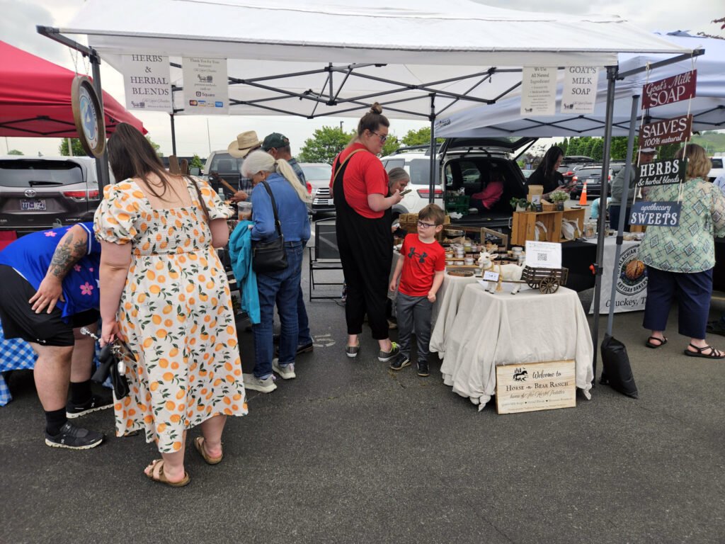 Outdoor market scene with shoppers under white tents browsing tables of goods and signs for herbs and soaps/treats. Horse and Bear Ranch