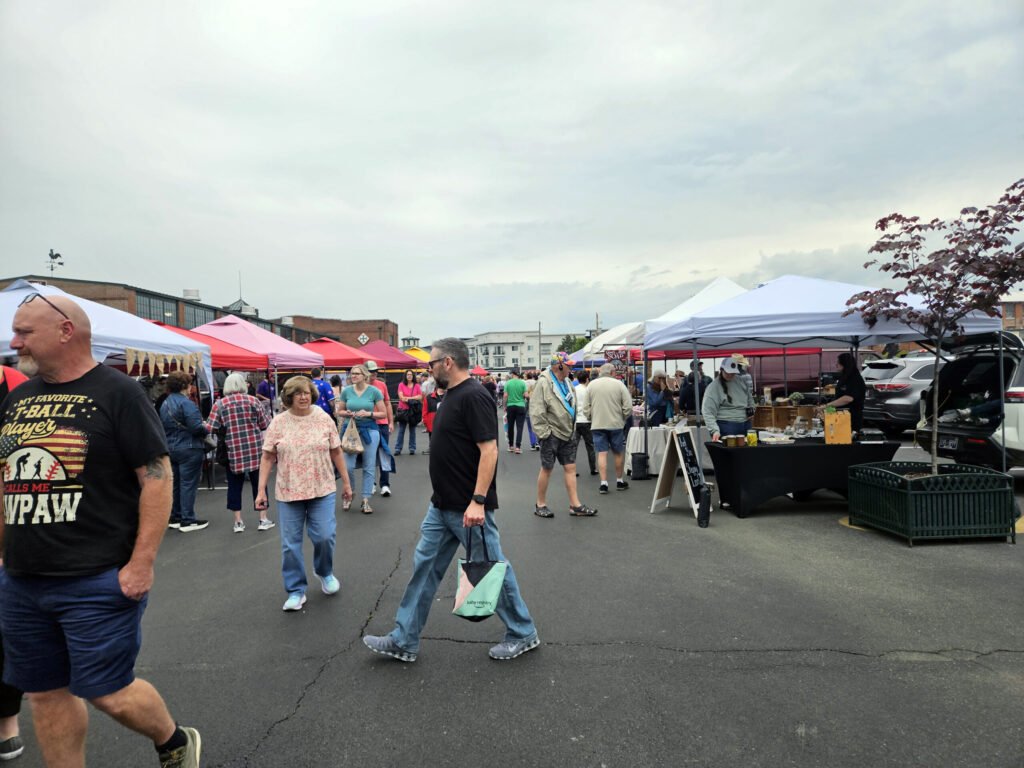 Outdoor market scene with colorful tents and shoppers walking between vendor stalls under a cloudy sky, urban buildings in the background.