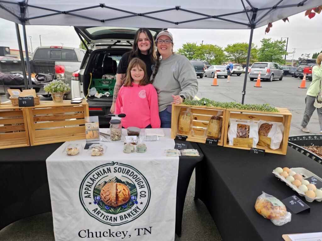 Smiling trio behind a market bakery stand, with loaves and pastries on display and a banner reading Appalachian Sourdough Co. Artisan Bakery (Chuckey, TN).