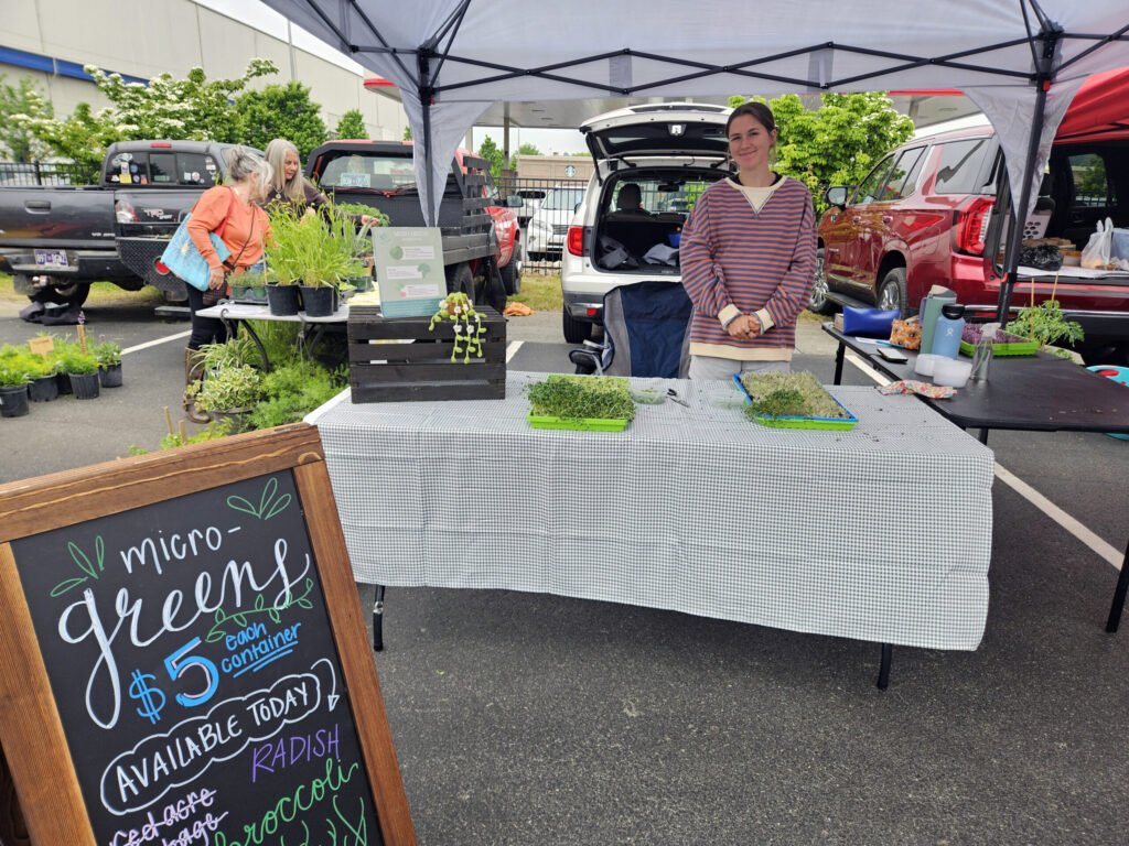 Market stall under a canopy selling microgreens; chalkboard sign and potted herbs on table, a smiling vendor in striped sweater behind the table.