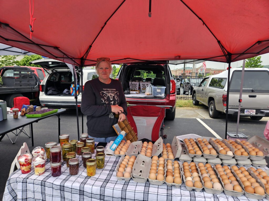 Woman standing behind a table under a red canopy at an outdoor market, with cartons of eggs and jars of jam on the table.