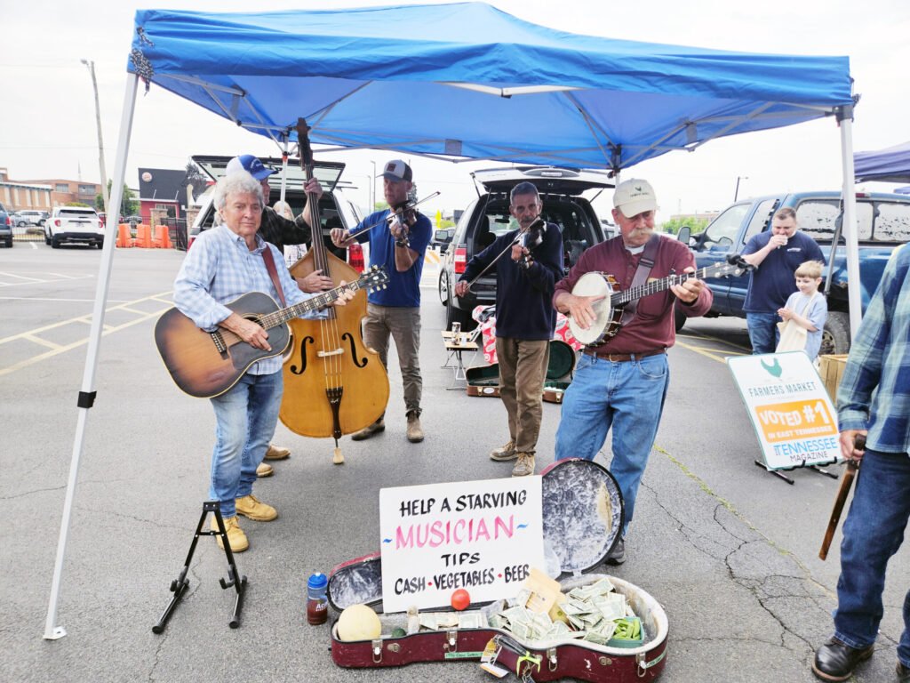 Street musicians perform under a bright blue canopy, playing guitar, upright bass, and violin to raise donations.