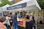 Outdoor market stall with a blue sign reading 'Cocoa Bean & Butter' under a white canopy; shoppers browse tables of small goods and samples.