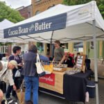 Outdoor market stall with a blue sign reading 'Cocoa Bean & Butter' under a white canopy; shoppers browse tables of small goods and samples.