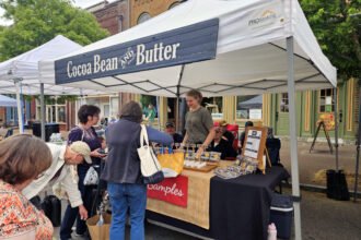 Outdoor market stall with a blue sign reading 'Cocoa Bean & Butter' under a white canopy; shoppers browse tables of small goods and samples.