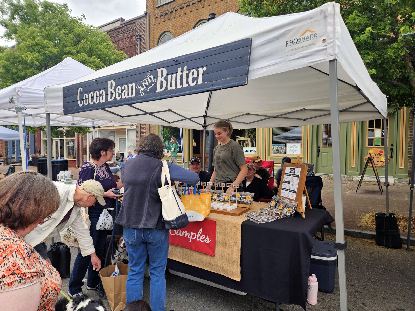 Outdoor market stall with a blue sign reading 'Cocoa Bean & Butter' under a white canopy; shoppers browse tables of small goods and samples.