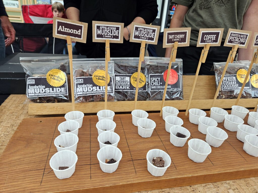 Assorted chocolate samples with flavor signs (Almond, Mudslide, Tart Cherry, Coffee, Pistachio) at a market booth on a wooden table, with white cups in front.