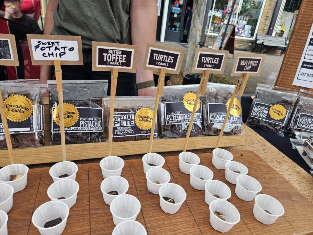 Assorted chocolate samples with flavor signs (Almond, Mudslide, Tart Cherry, Coffee, Pistachio) at a market booth on a wooden table, with white cups in front.