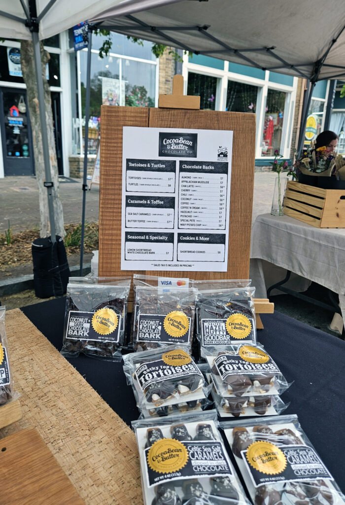 Outdoor market stall selling Cocoa Bean & Butter chocolates on a wooden display board with packaged chocolate bars in front