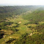 View of a valley with patchwork fields, scattered farmhouses, and forested hills.
