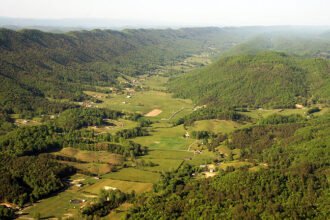 View of a valley with patchwork fields, scattered farmhouses, and forested hills.