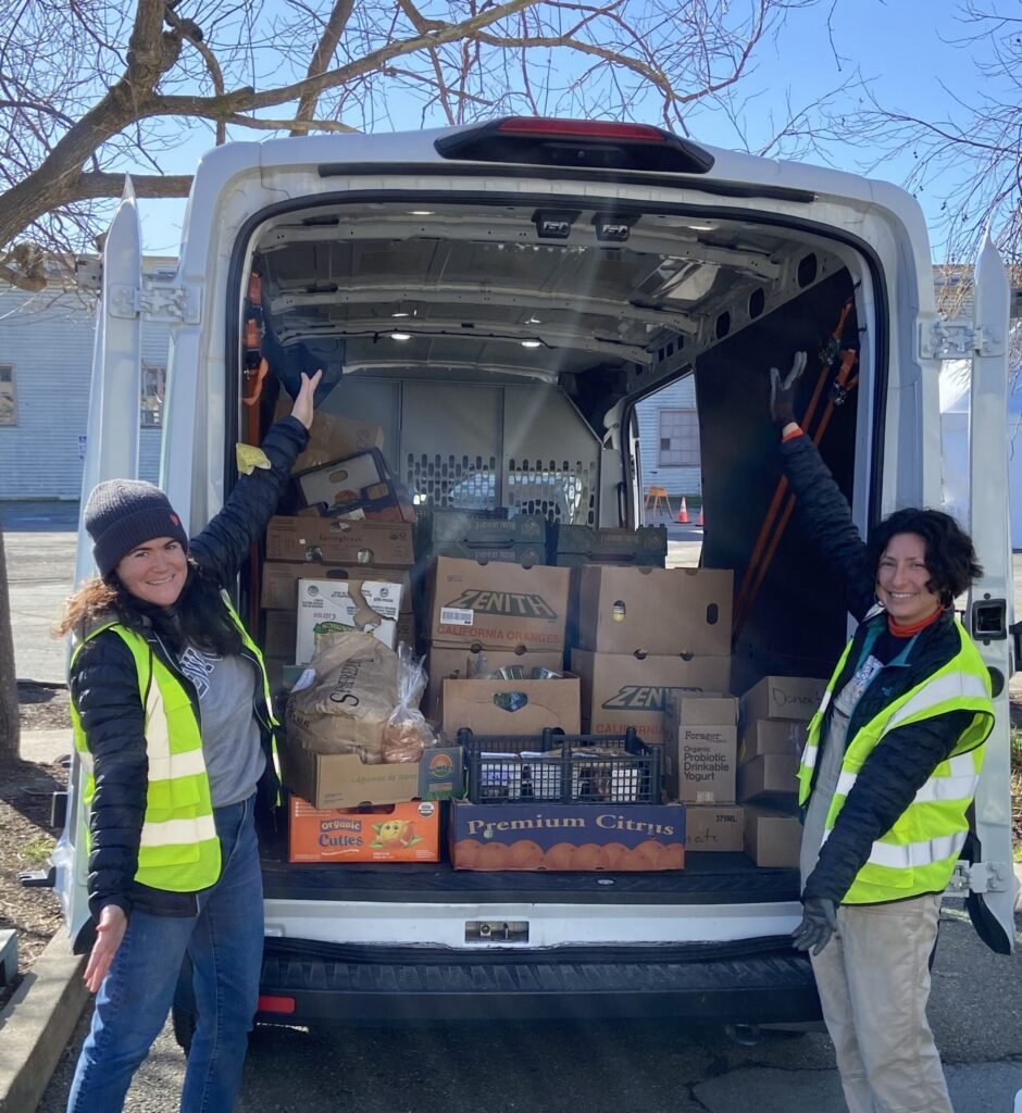 People in high-visibility vests stand at an open van filled with stacked cardboard boxes and bags of groceries; two individuals reach inside to unload.