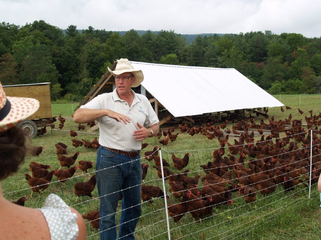 Joel Salatin explains farm practices to visitors near a large coop with many brown chickens inside a fenced yard.  - Photo by Nick V from Washington dc, CC BY-SA 2.0 Via Wikimedia Commons