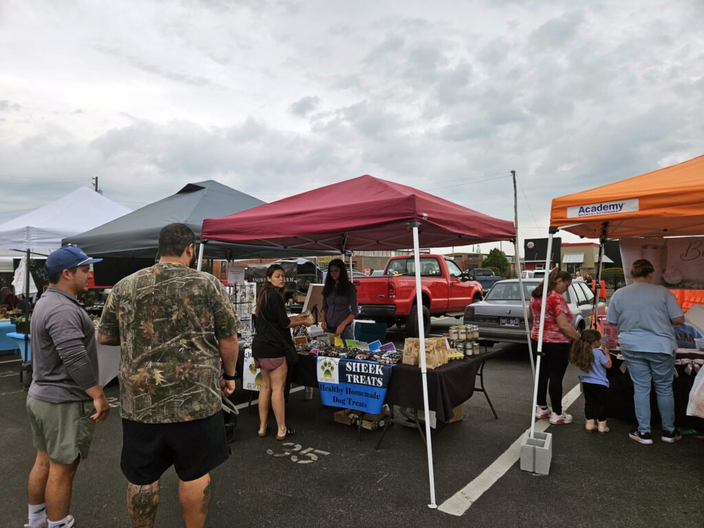 Outdoor market scene with white, gray, and red tents and shoppers browsing vendor tables under a cloudy sky with parked cars in the background.