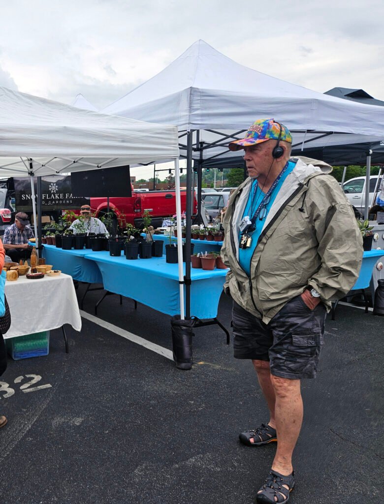 Older man wearing a colorful tie-dye cap and headset stands near vendor tables at an outdoor market under white tents.
