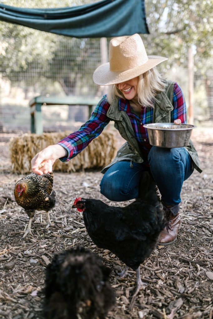 Woman kneels in a farm yard, wearing a wide-brimmed hat and plaid shirt, feeding chickens with a bowl nearby.