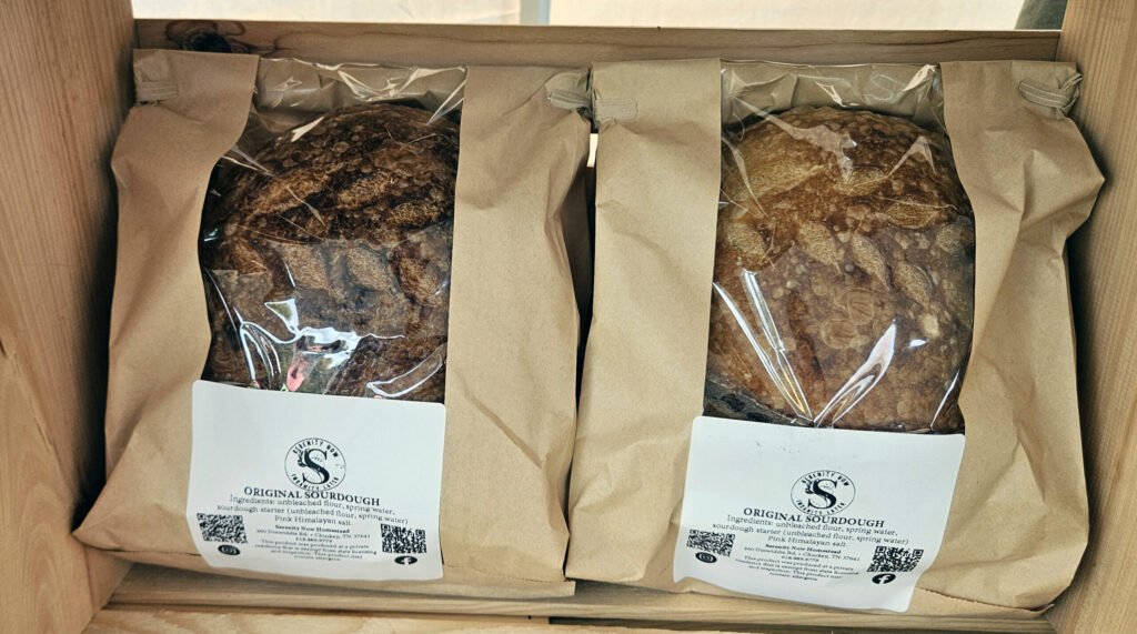 Two loaves of original sourdough in brown paper bags with clear plastic windows, placed in a wooden crate with white labels showing ingredients and branding beneath the bags.