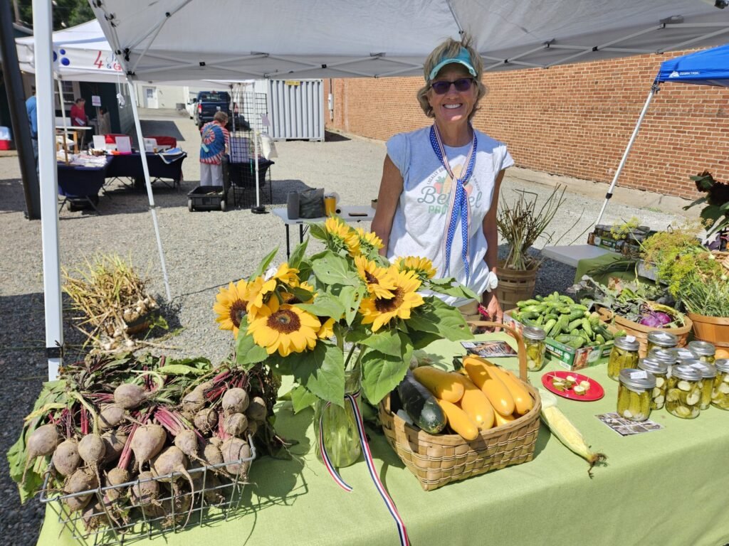 Smiling woman at a vendor table with sunflowers and fresh produce at an outdoor market under white canopies.