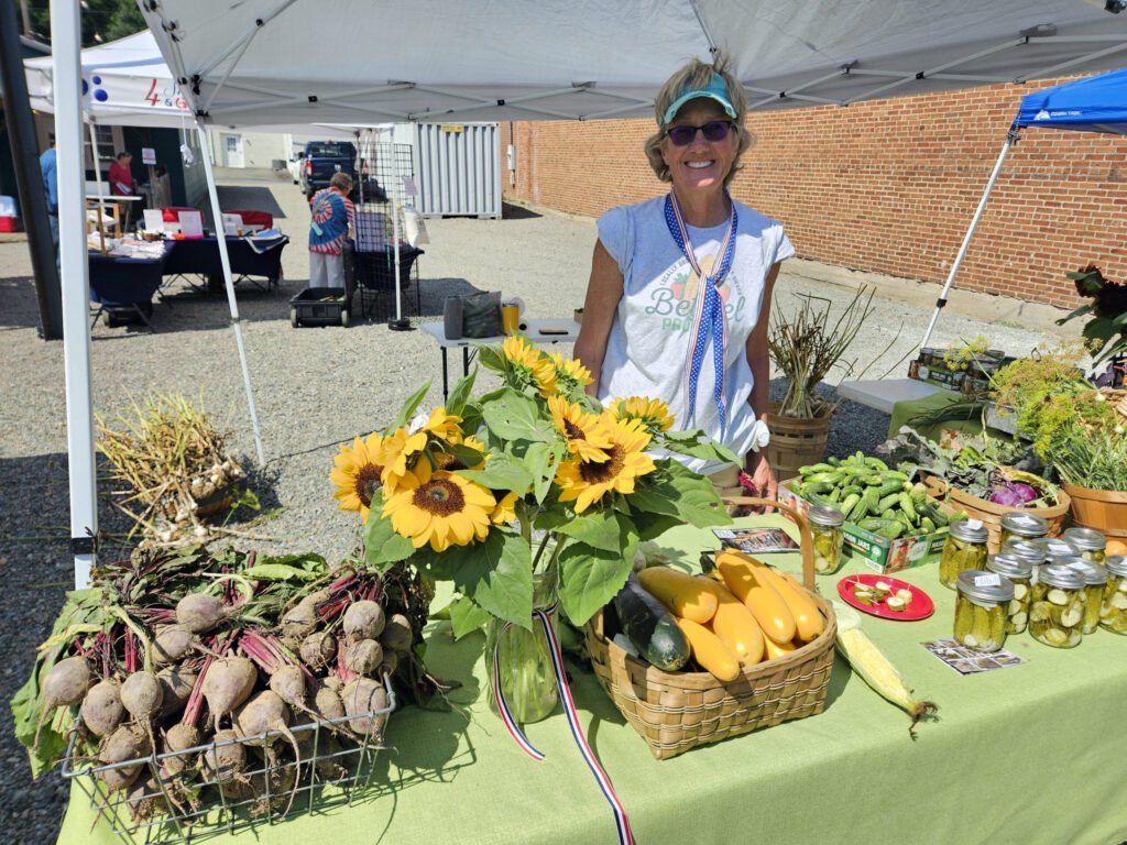 Vendor at an outdoor market smiling behind a display of sunflowers, squash, and jars of preserved goods under a white canopy.