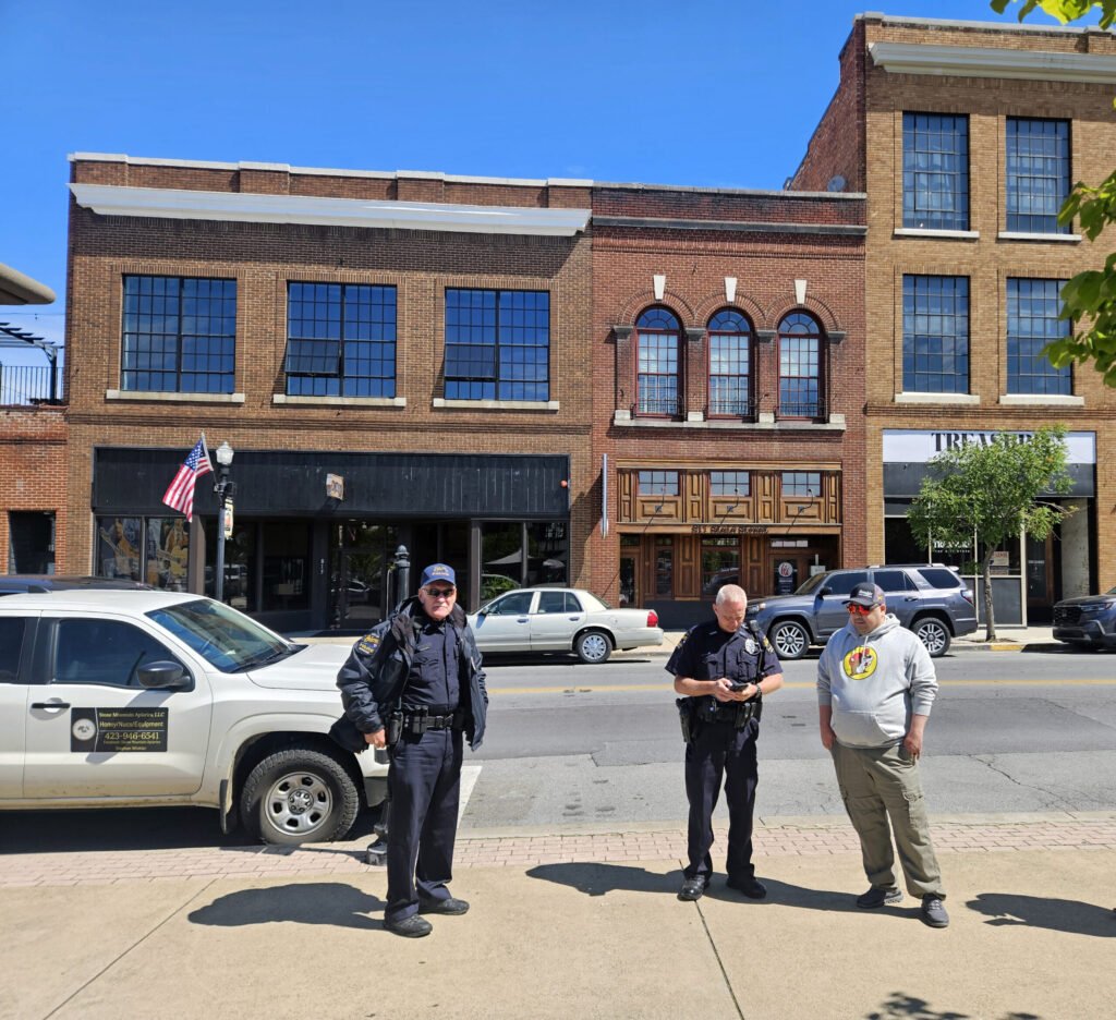 Two police officers stand on a city street beside a man in a hoodie in front of a brick building with large storefront windows and parked cars behind them, an American flag nearby