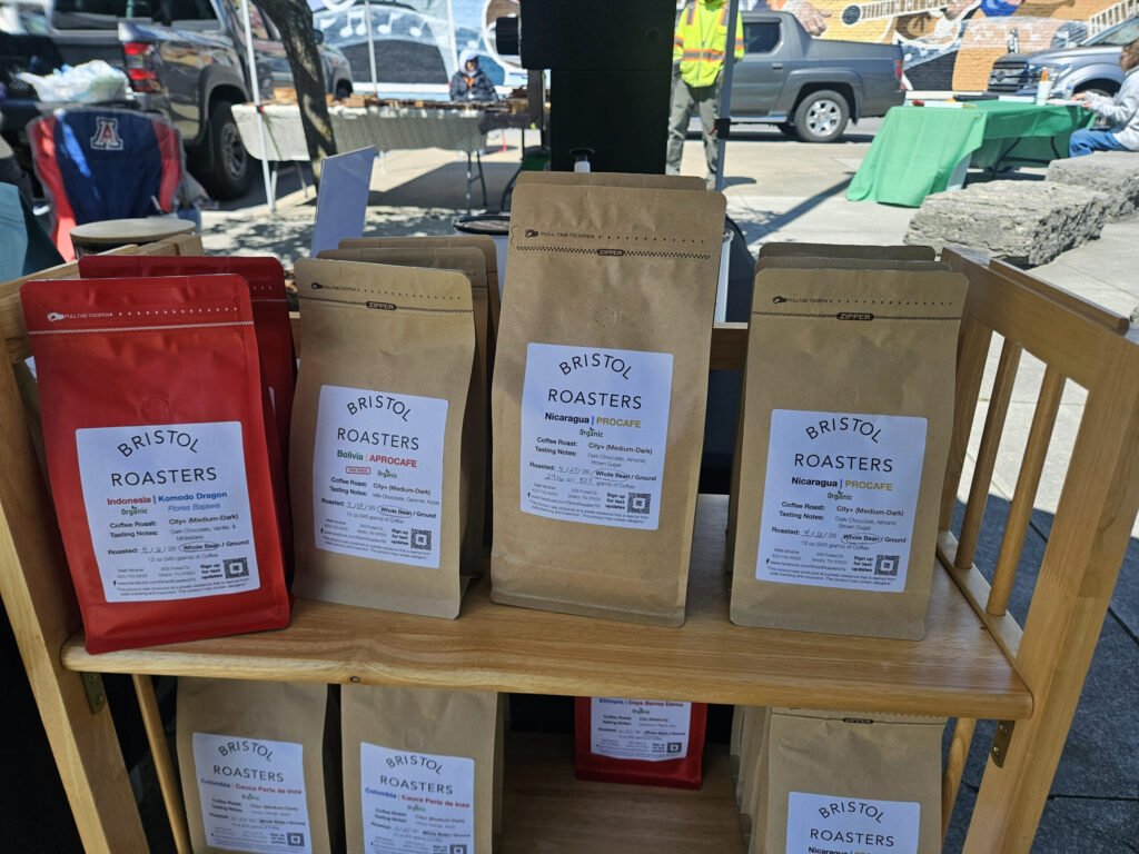 Brown and red coffee bags labeled Bristol Roasters arranged on a wooden shelf at an outdoor market.