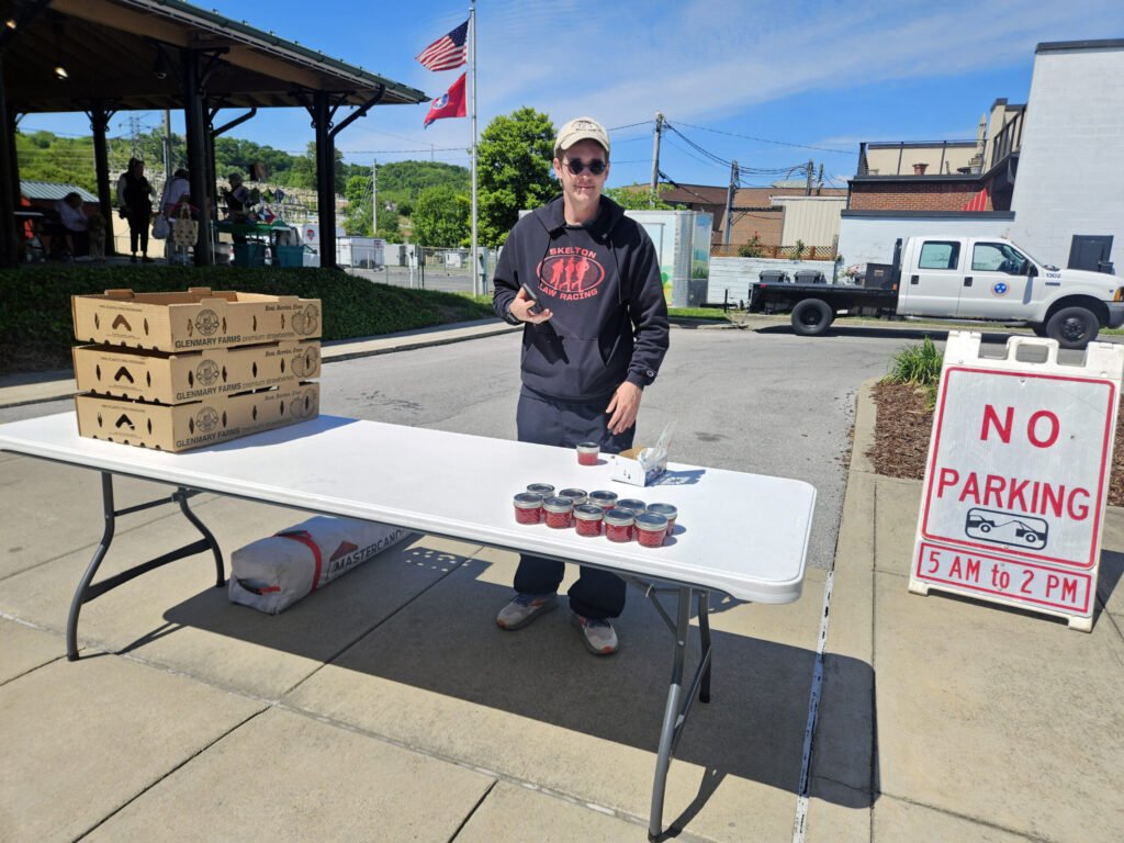 Man in a dark hoodie and cap stands behind a table with small red jars, at an outdoor market or community event.