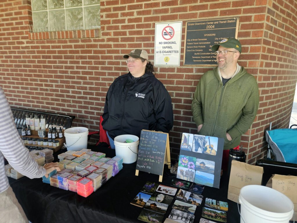 Two people stand behind a soap stall at a brick wall, with colorful soaps and postcards on display.