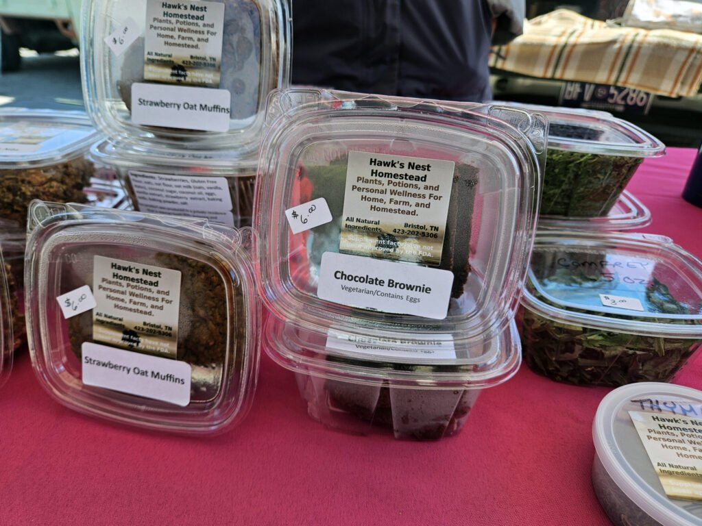 Plastic containers on a pink table labeled Hawk's Nest Homestead bake goods, including Strawberry Oat Muffins and Chocolate Brownie.