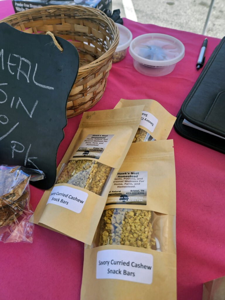 Close-up of a market table with brown snack-bar bags labeled 'Savory Curried Cashew Snack Bars' beside a wicker basket on a pink tablecloth.