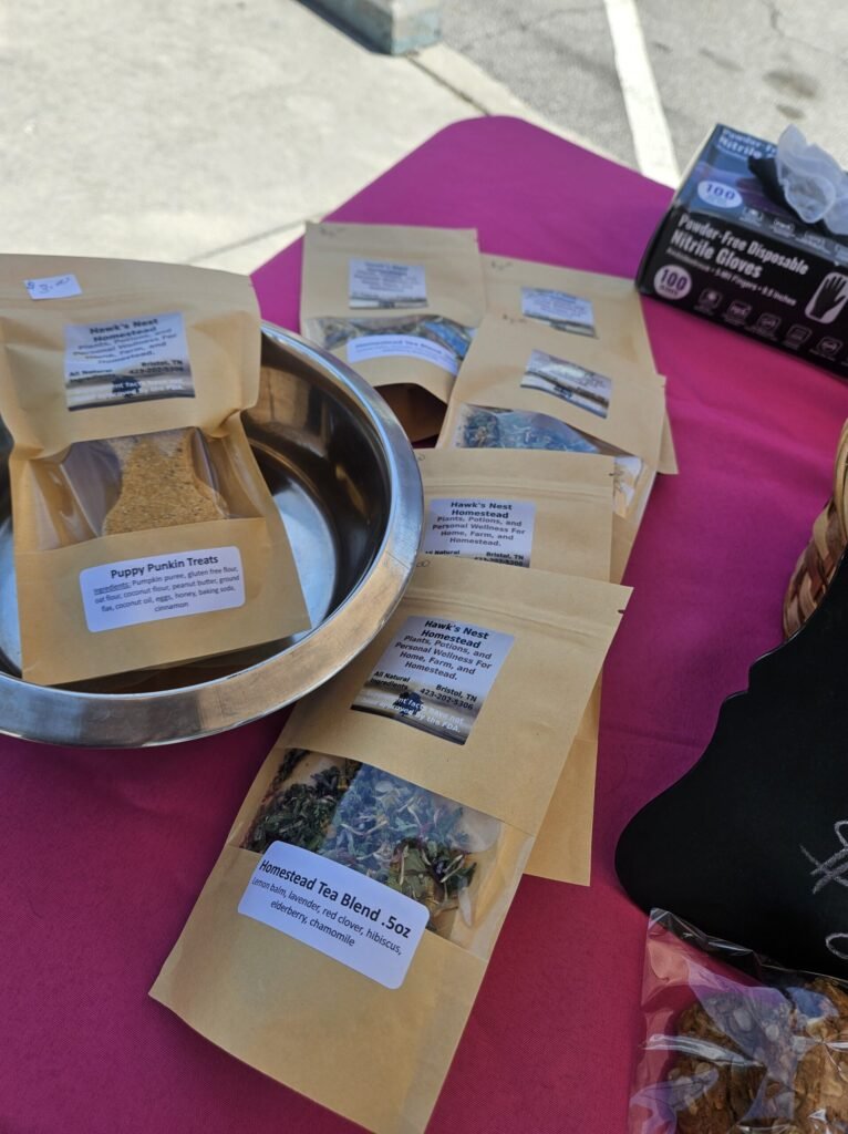 Kraft tea bags labeled 'Hawk's Nest Homestead' spread on a pink table with a metal bowl and a box of disposable gloves nearby