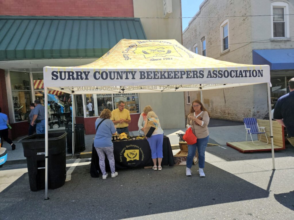 Yellow canopy tent for Surry County Beekeepers Association at an outdoor street fair, with people browsing tables of beekeeping gear and informational materials.