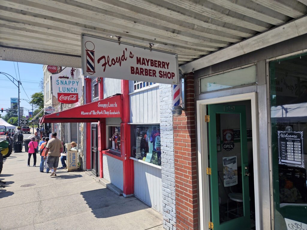 Street sidewalk with storefronts and vintage signage including Floy d's Mayberry Barber Shop and Snappy Lunch; pedestrians walk by on a sunny day (urban neighborhood).