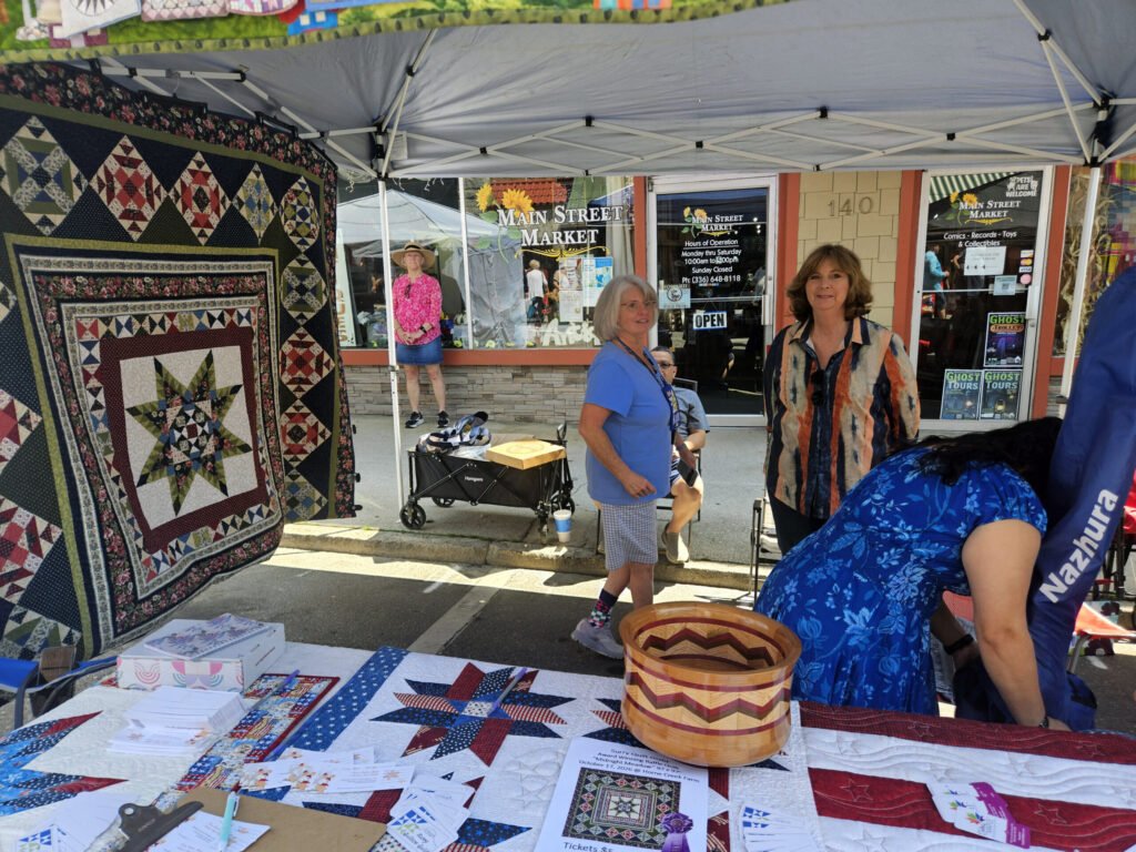 Quilt stall at an outdoor market with patchwork quilts on display, baskets, and printed brochures; two women nearby under a canopy.
