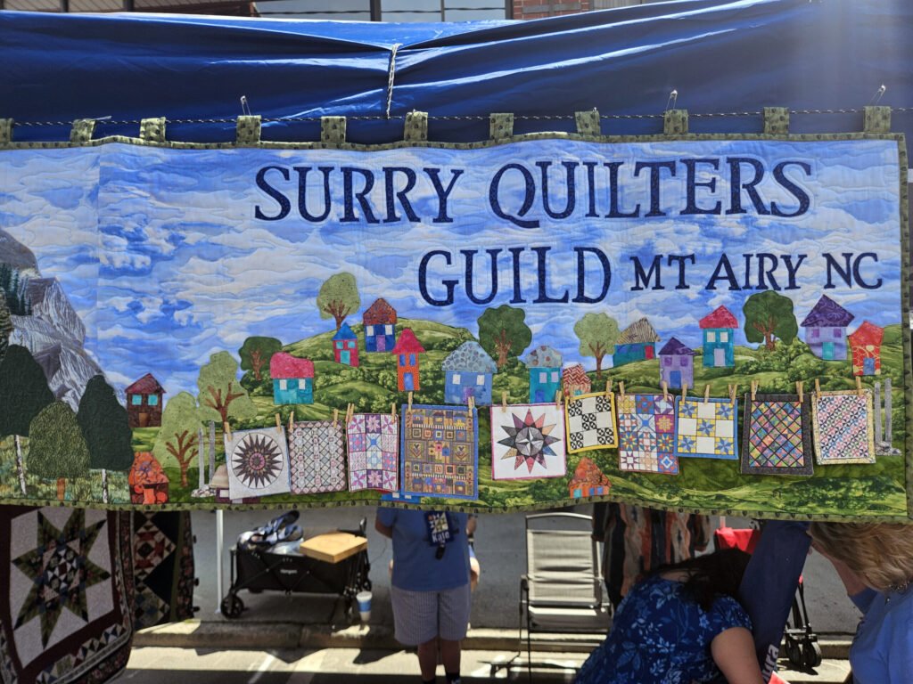 Banner reading'Surry Quilters Guild Mt Airy NC' displayed at an outdoor booth, with colorful quilt houses in the scene.
