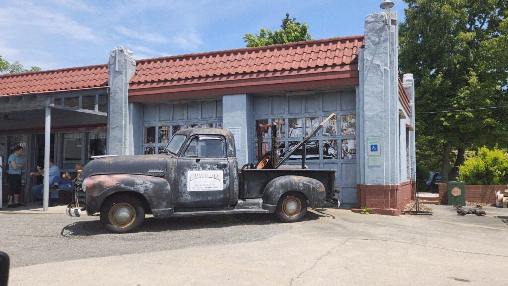 Old rusty pickup truck parked in front of a blue-gray storefront with a red tile roof; people sit to the left.